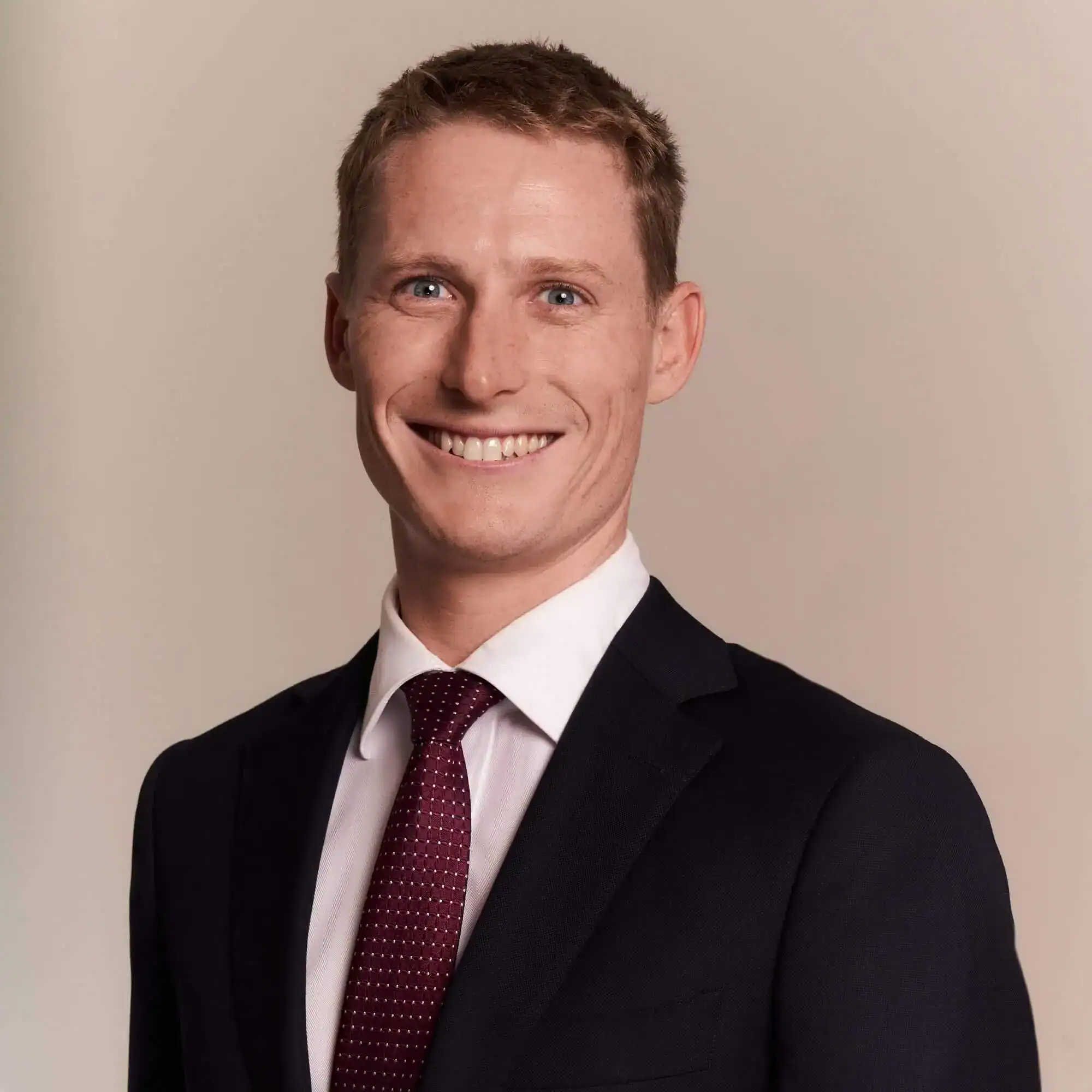 A man in a suit and tie smiles at the camera against a plain light background.
