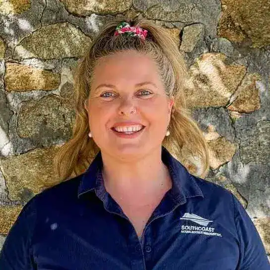 Smiling woman with blond hair in a ponytail stands in front of a stone wall, wearing a navy shirt.