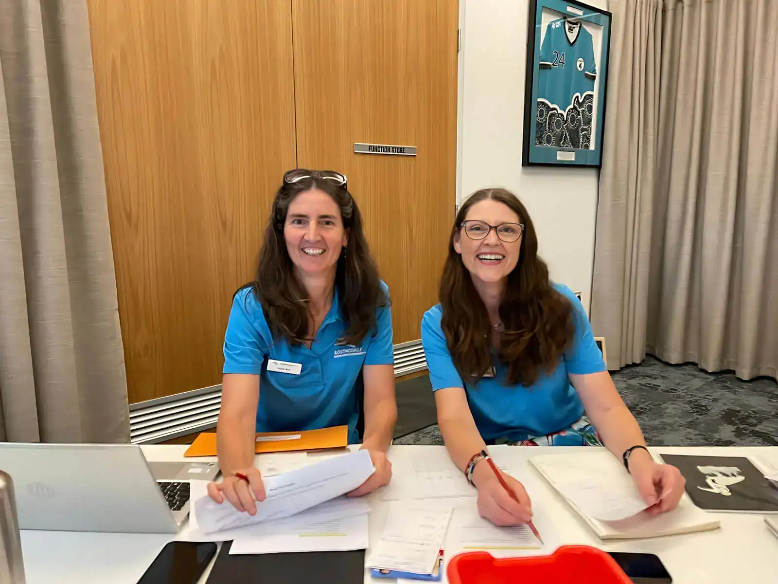 Two women in blue shirts smile while working at a table with papers and a laptop.