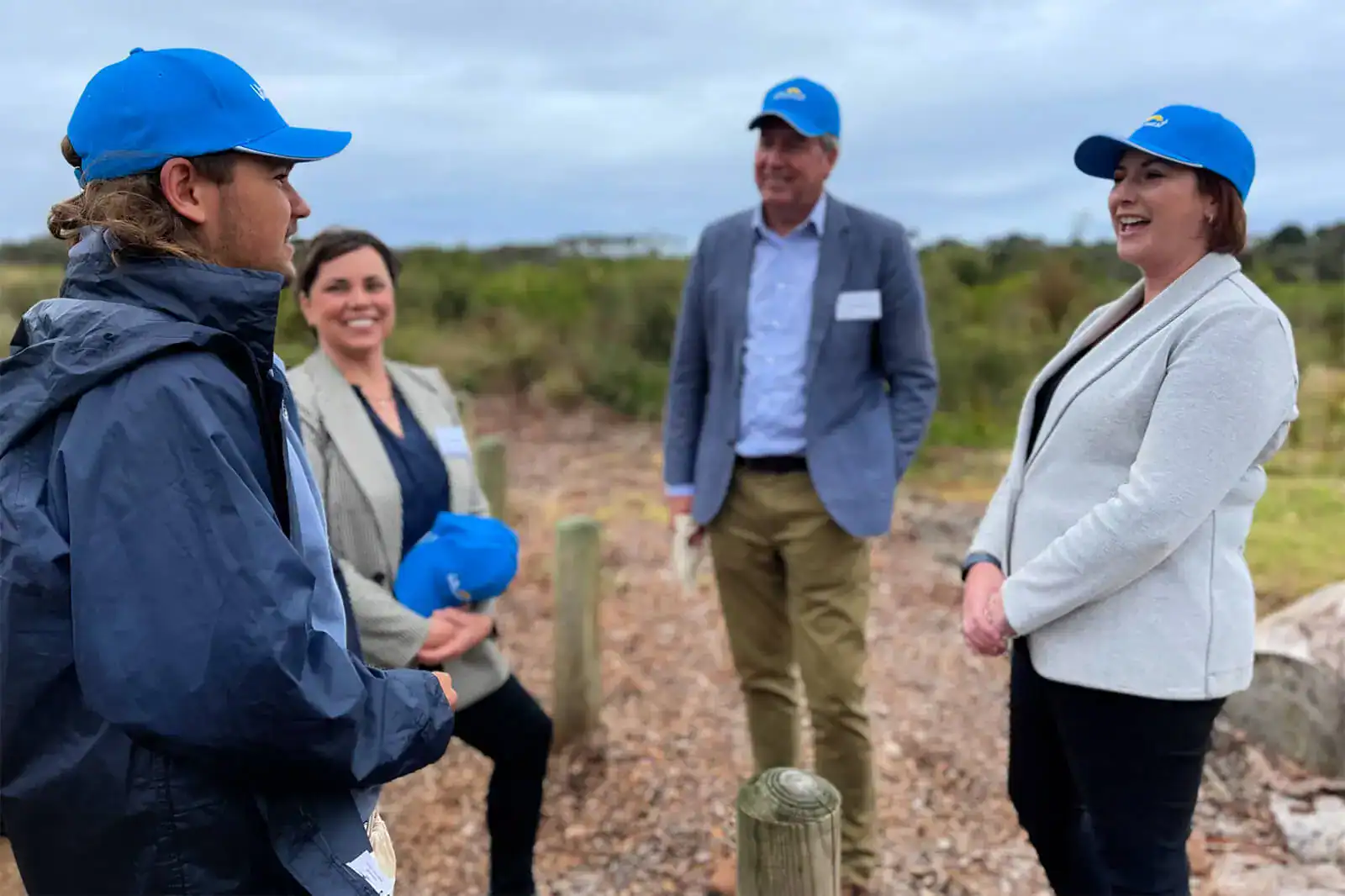 Four people wearing blue caps chat outdoors on a cloudy day, smiling and standing on a gravel path.