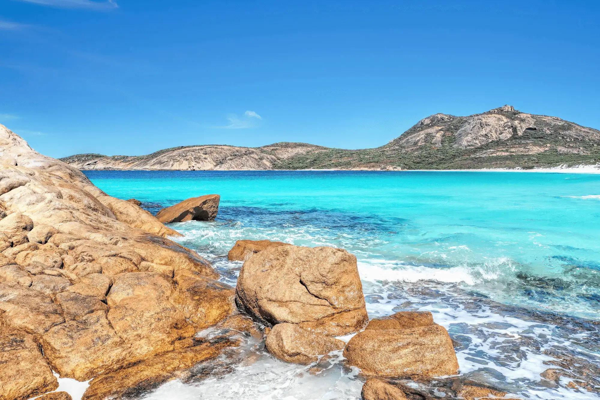 Rocky shoreline with turquoise water and distant hills under a clear blue sky.