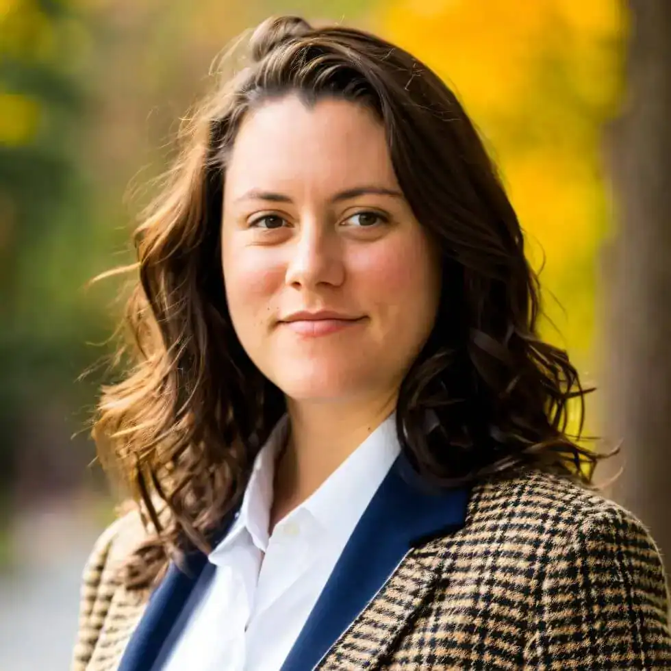 A woman with wavy brown hair in a checked blazer stands outdoors with a blurred autumn background.