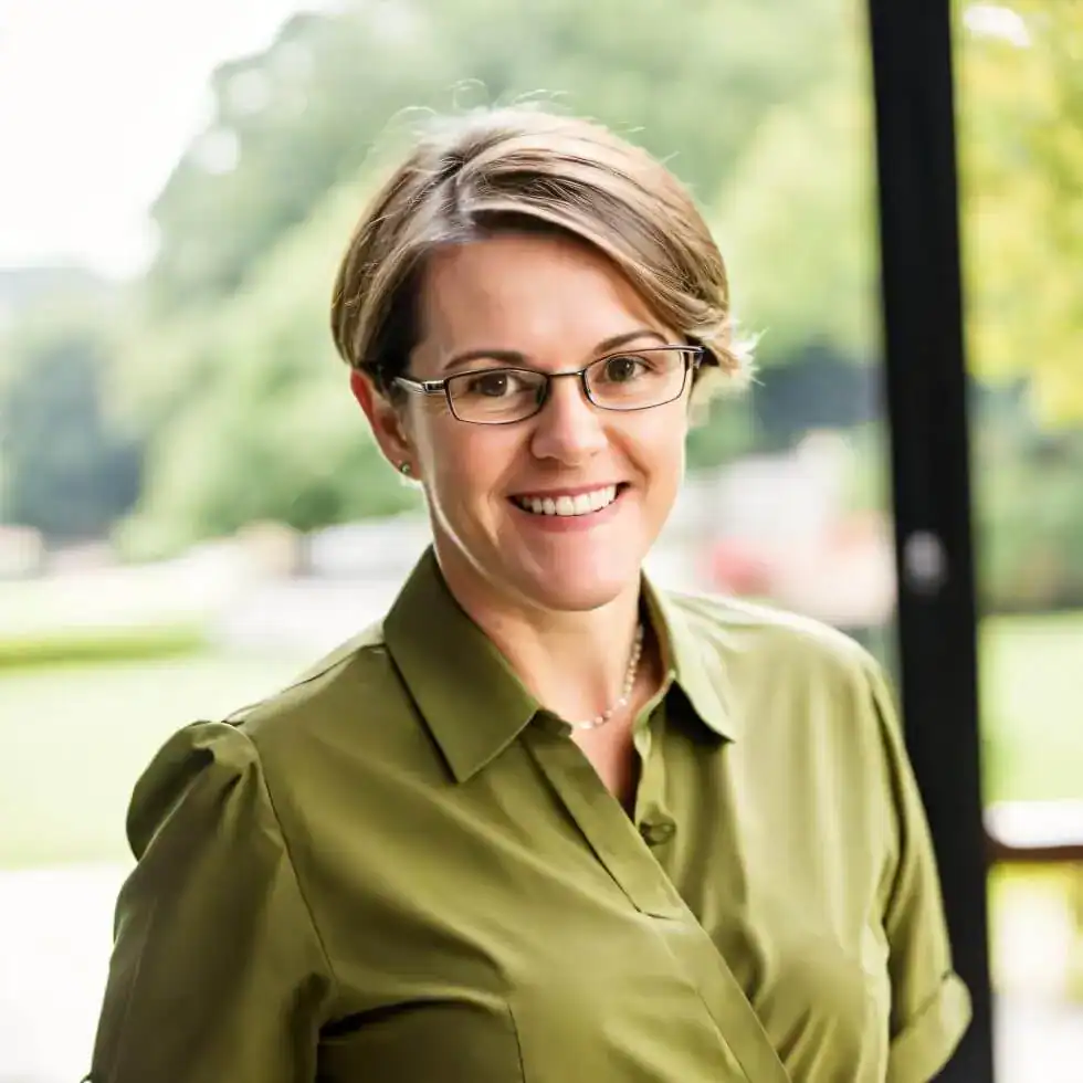 Smiling woman with short brown hair and glasses, wearing a green shirt, standing outdoors.