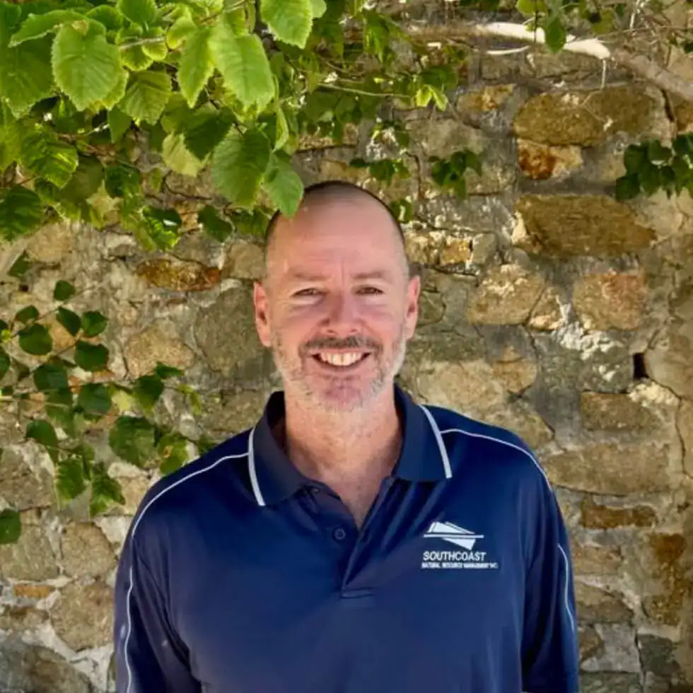 Smiling man in a blue polo shirt stands in front of a stone wall with green leaves overhead.