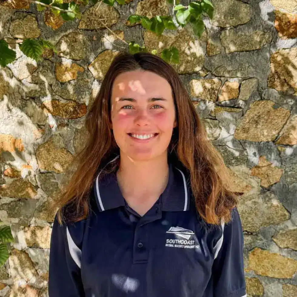 Woman with long brown hair smiling, standing outside against a stone wall, wearing a navy polo shirt.