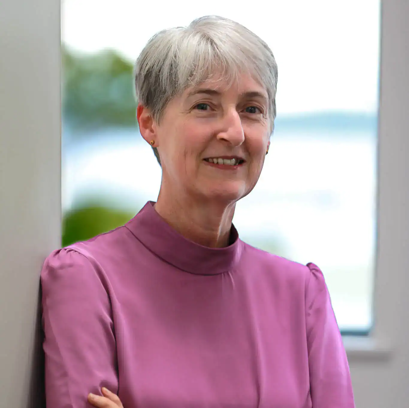 Smiling older woman with short gray hair, wearing a purple blouse, stands indoors with arms crossed.
