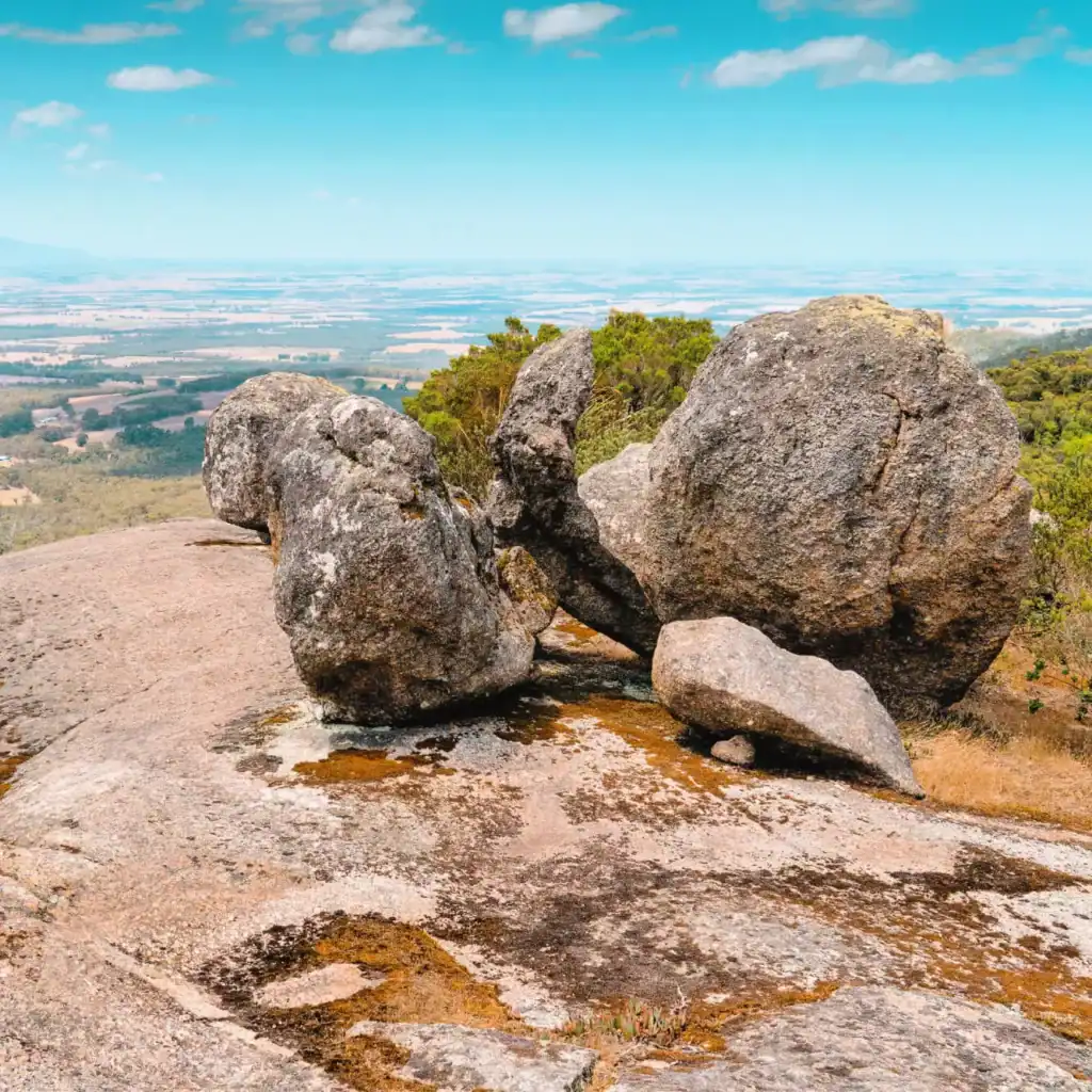 Large granite boulders on a rocky hilltop overlook a scenic, distant landscape under a bright blue sky.