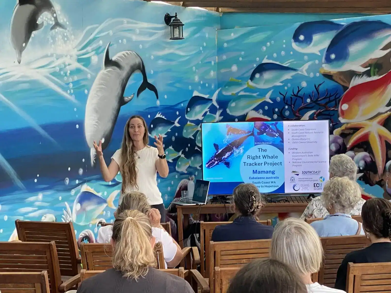 A woman presents to a seated audience about whale tracking, with ocean-themed murals and slides behind her.
