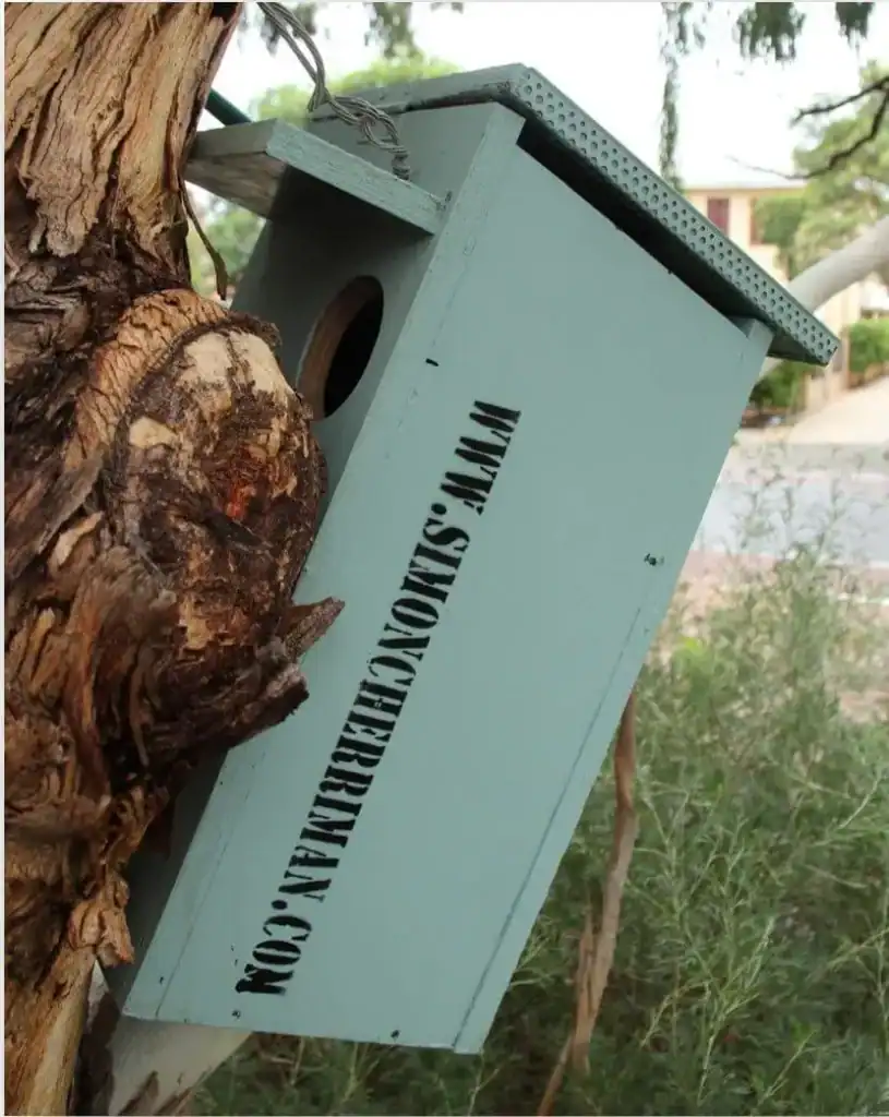 A blue birdhouse attached to a tree trunk with "www.simoncheridan.com" written on its side.