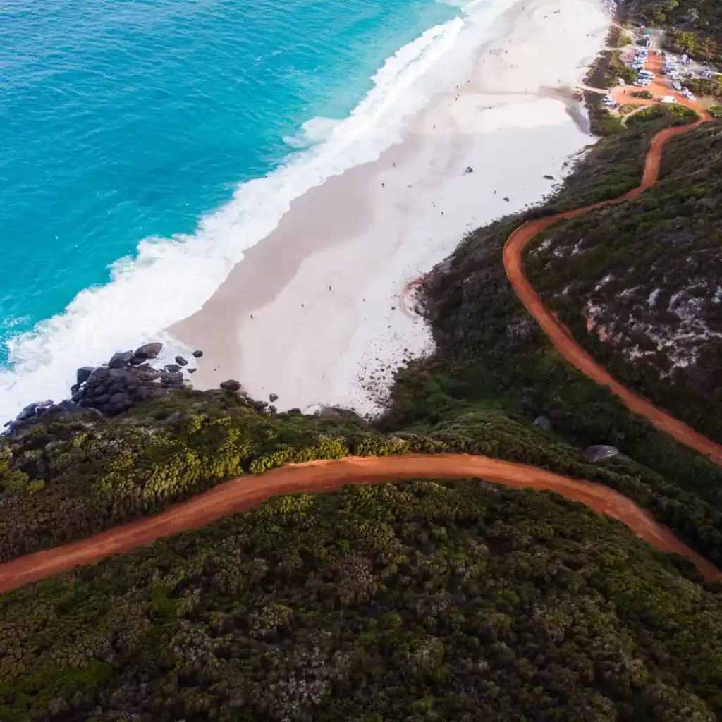 A winding red dirt road runs along green cliffs above a white sandy beach and turquoise ocean waves.