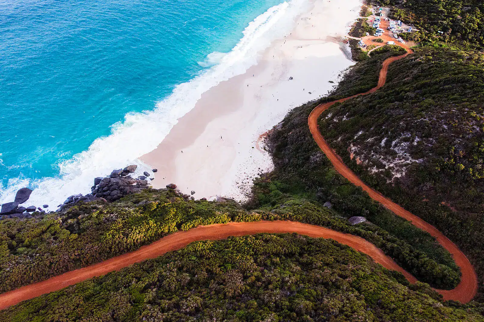 A winding red dirt road runs along a lush green hillside next to a white sandy beach and bright blue ocean.