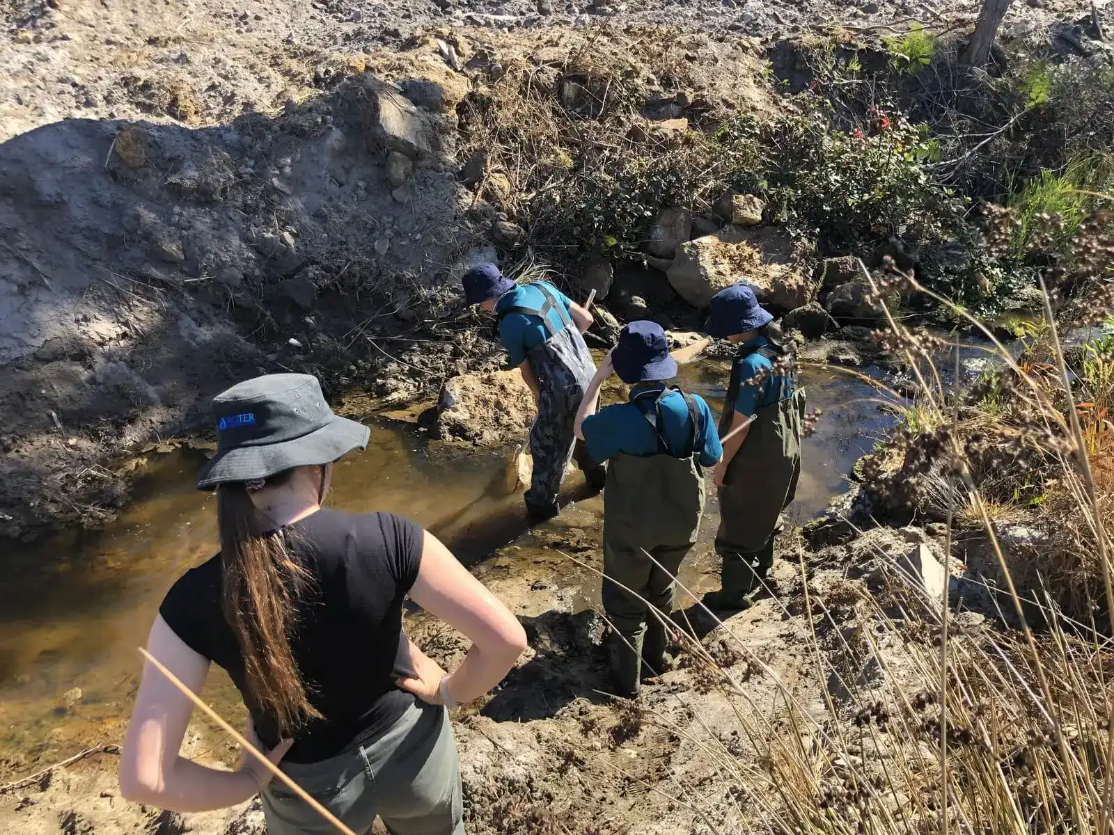 Four people in outdoor gear stand by a shallow stream, observing the water and surroundings on a sunny day.