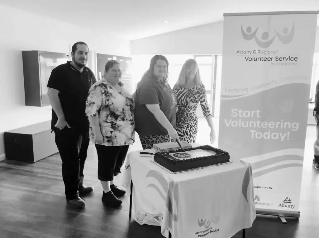 Four people stand behind a table with a cake and a "Start Volunteering Today!" banner in the background.