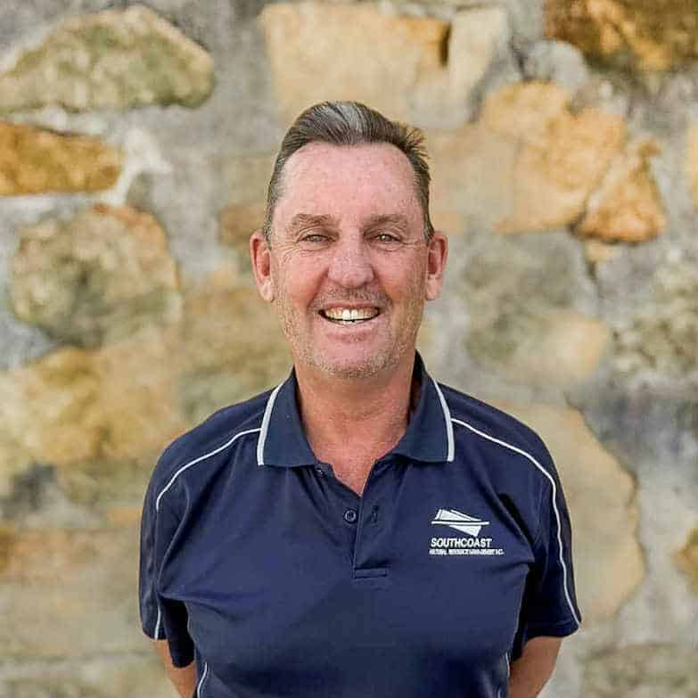 Smiling man in a navy shirt stands in front of a stone wall.