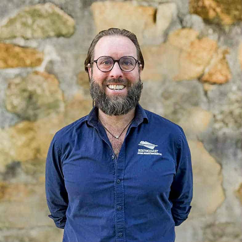 Man with glasses and a beard, wearing a navy shirt, smiling in front of a stone wall.