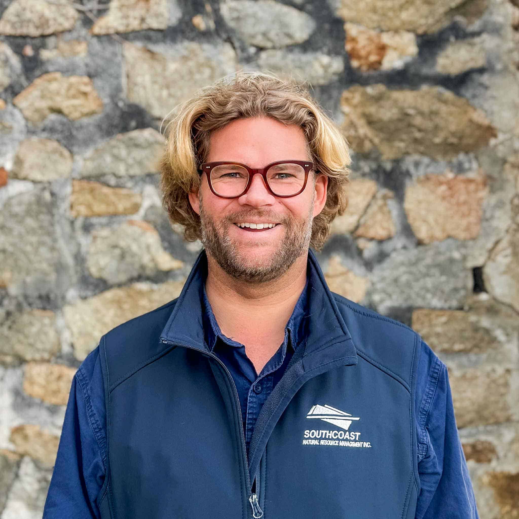 Smiling man with glasses and blond hair stands in front of a stone wall, wearing a navy vest and shirt.