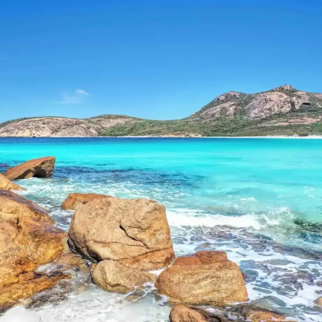 Clear turquoise water, large rocks, and green hills under a bright blue sky at a scenic coastline.