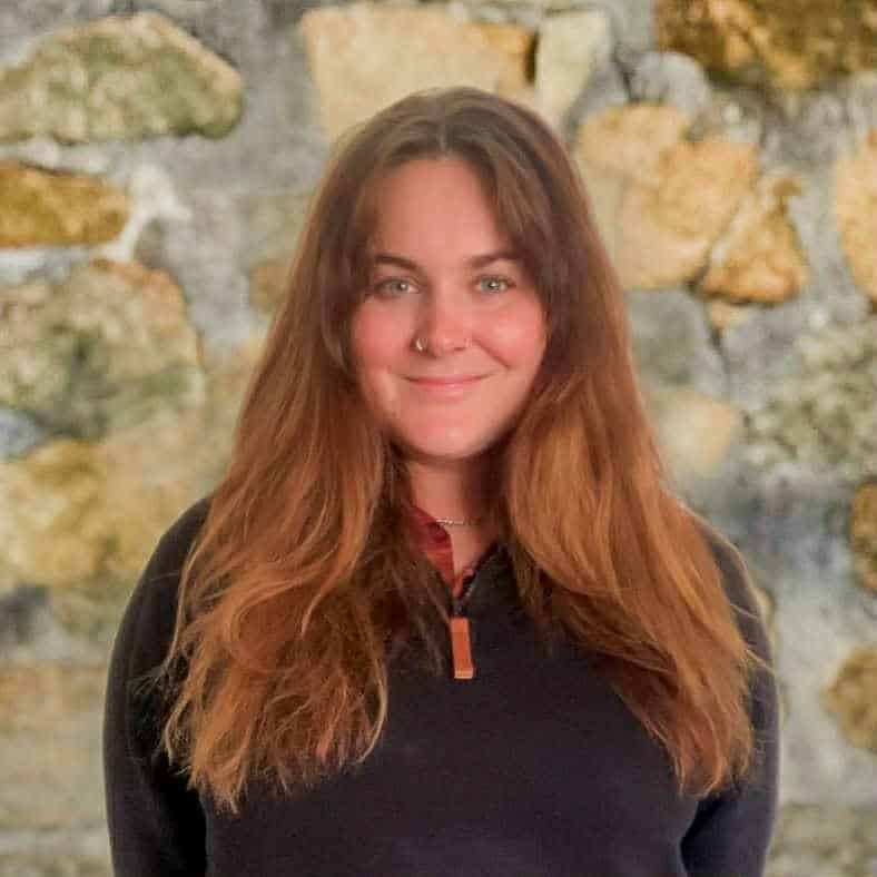 Woman with long brown hair and a nose ring, smiling in front of a stone wall background.