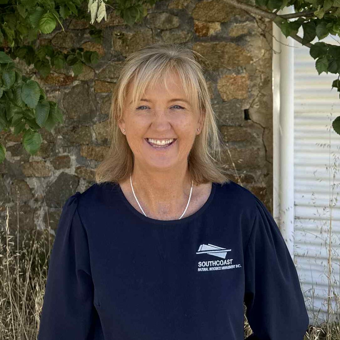 Smiling woman in a navy shirt stands outdoors by a leafy tree and stone wall, with dry grass in the foreground.