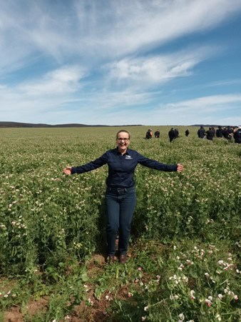 Person standing with arms outstretched in a field of green plants with groups of people in the background.