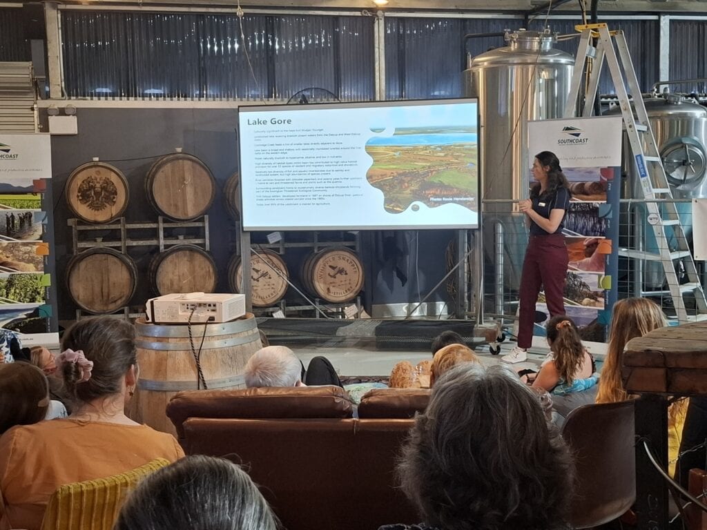 A woman presents a slideshow to a seated audience in a brewery with barrels and metal tanks around.