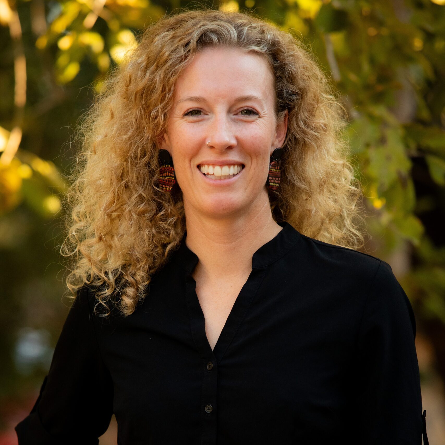 Smiling woman with curly blonde hair, wearing a black shirt and yellow pants, standing outdoors.
