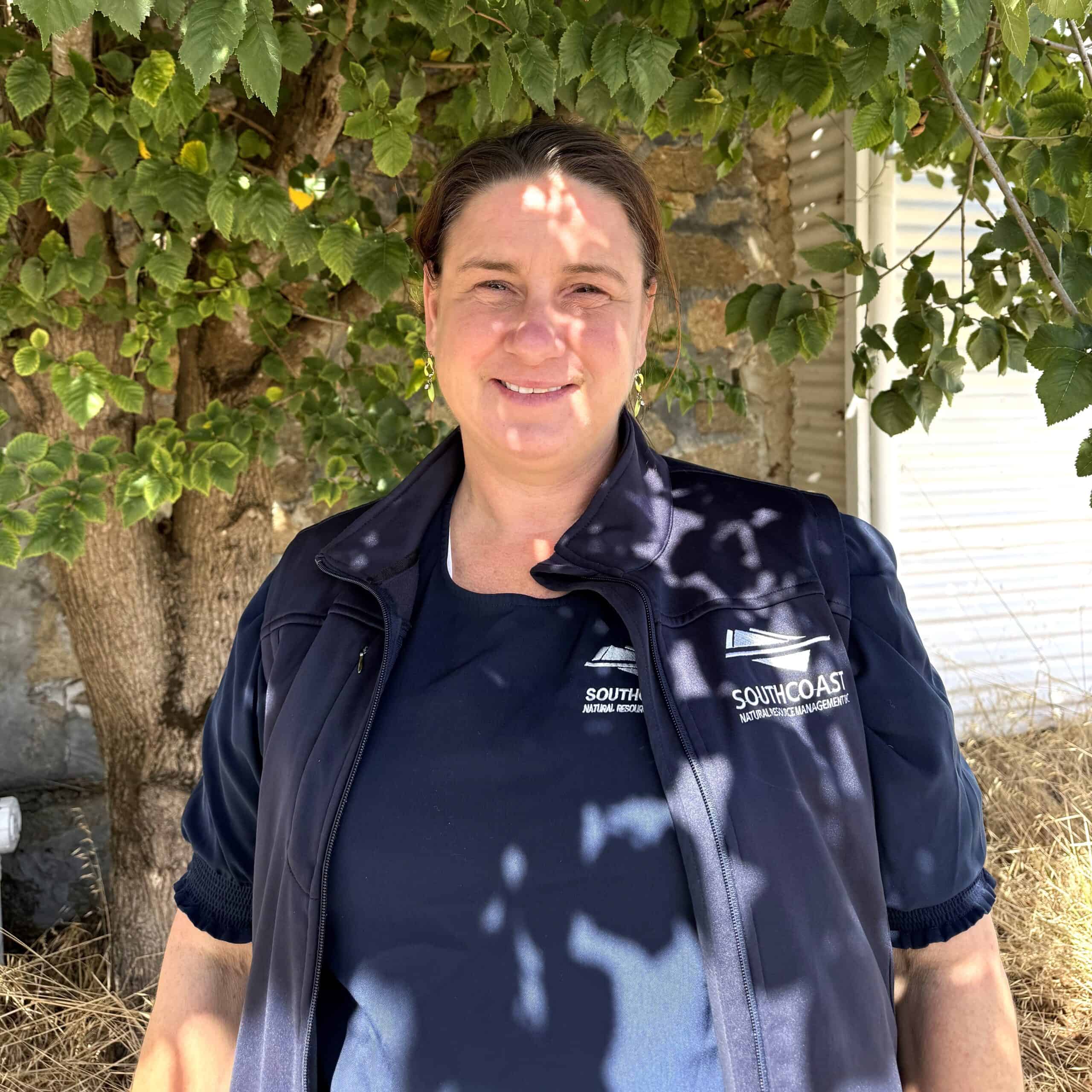 Woman in a navy waistcoat stands outside under a leafy tree, smiling at the camera in bright sunlight.