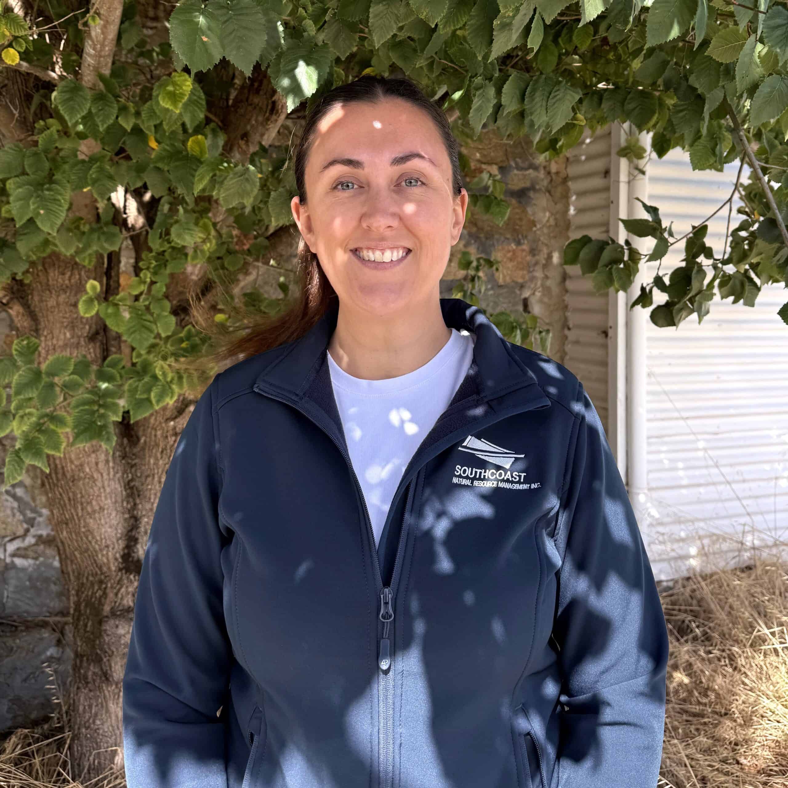 Woman smiling under a leafy tree, wearing a dark jacket with "Southcoast" text on it.