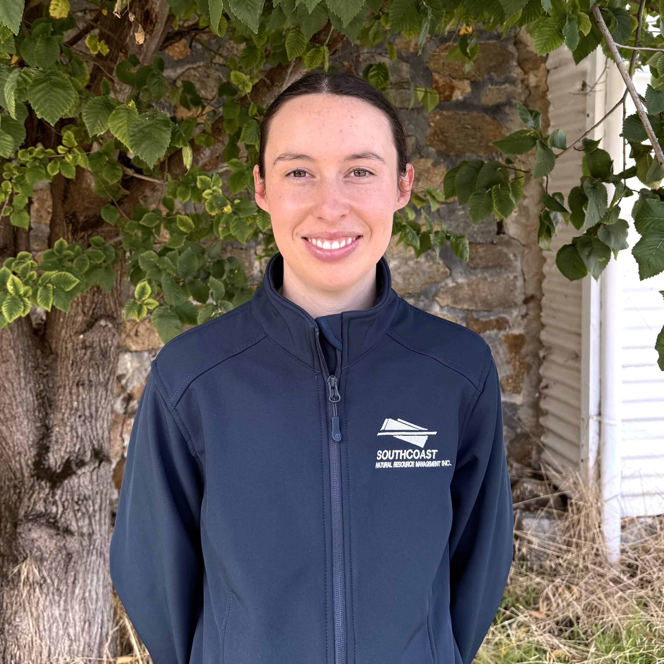 A woman in a navy jacket stands smiling in front of a leafy tree and a stone wall.