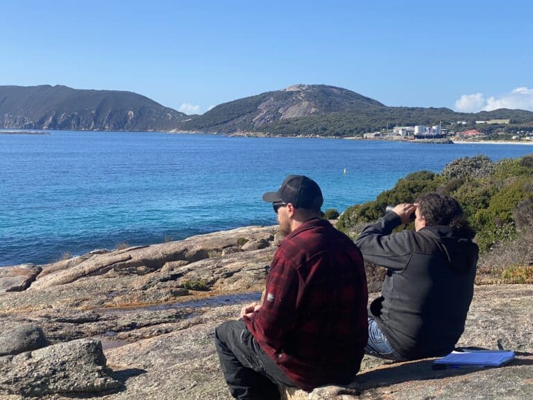 Two people sit on rocks by a blue sea, looking toward distant hills under a clear, sunny sky.