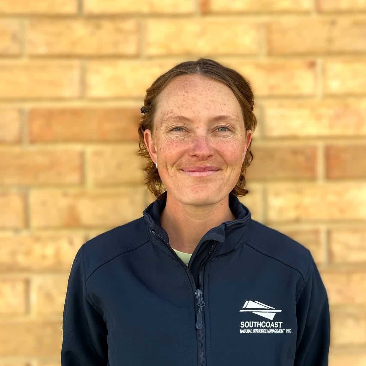 Person in a navy Southcoast jacket stands smiling in front of a tan brick wall.