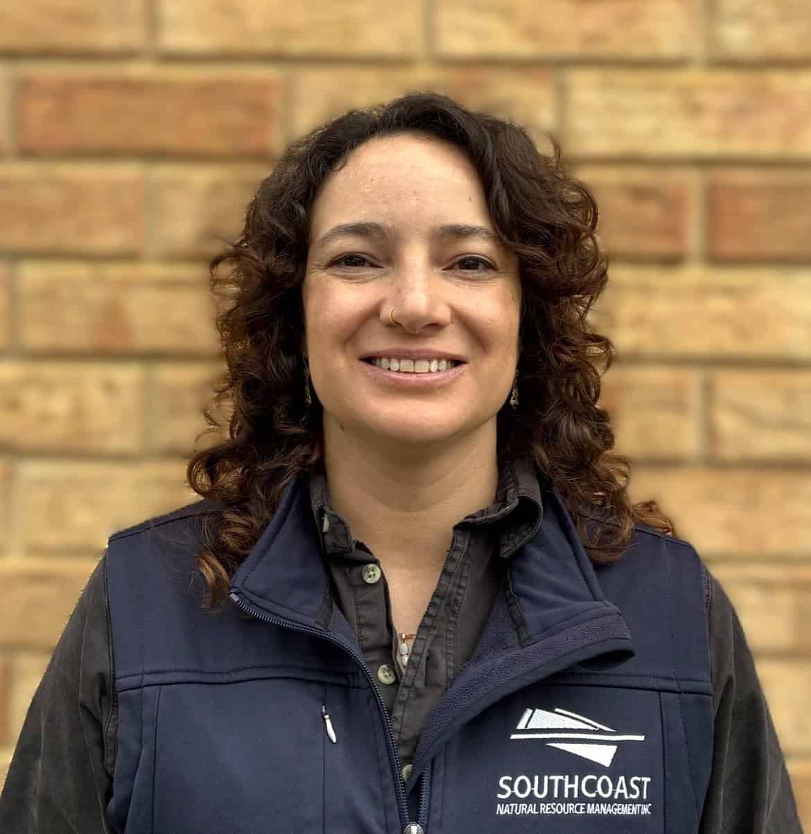 Woman with curly hair smiling, wearing a navy vest with “Southcoast Natural Resource Management” logo.