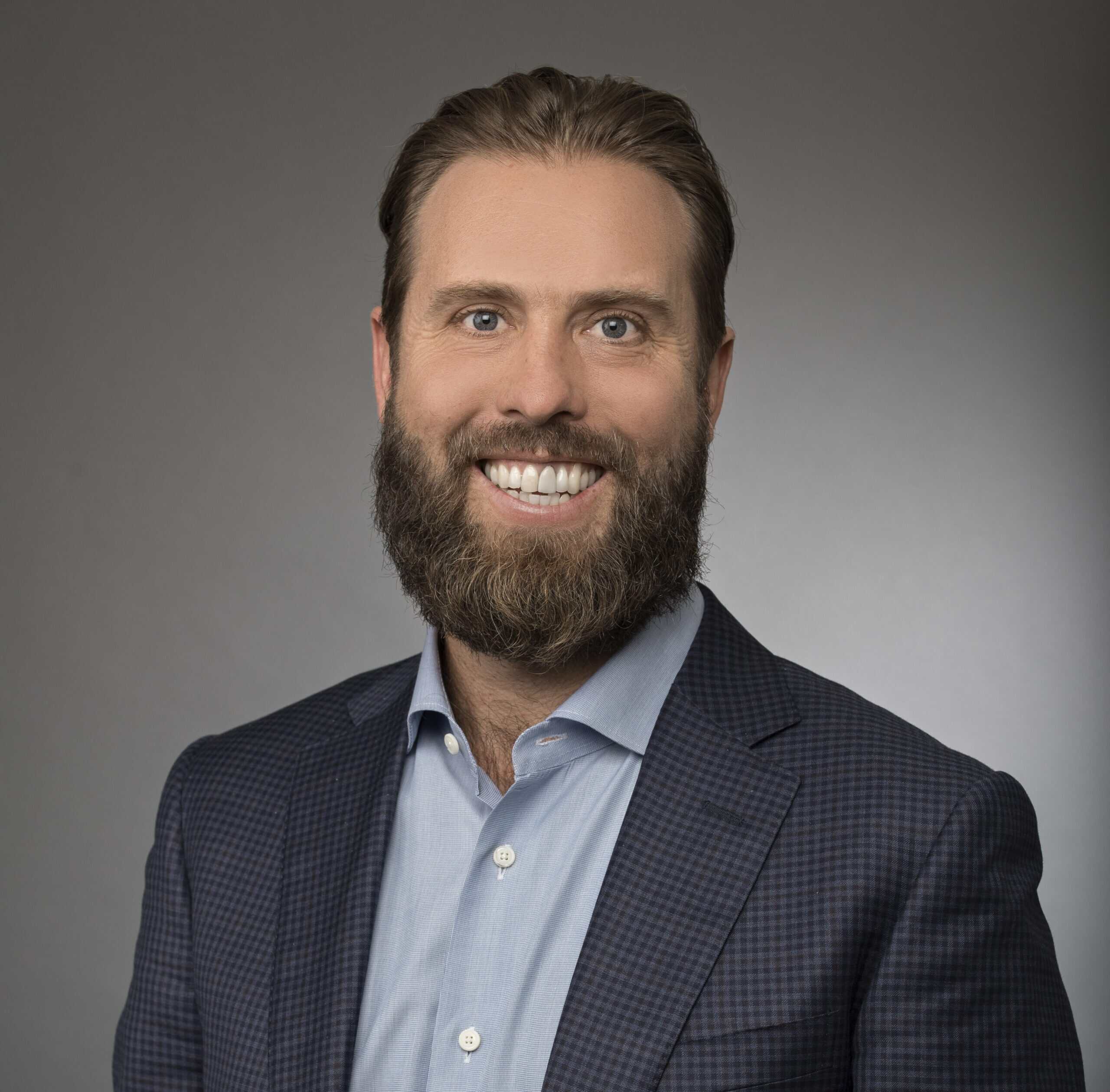 Smiling man with a beard in a blue suit and light blue shirt, posing against a gray background.