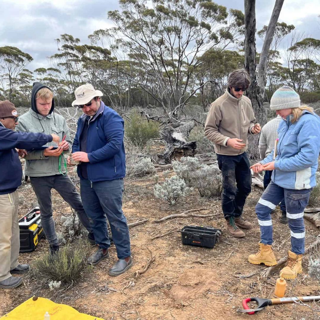 Five people stand outdoors in the bush, handling equipment and tools, with trees in the background.