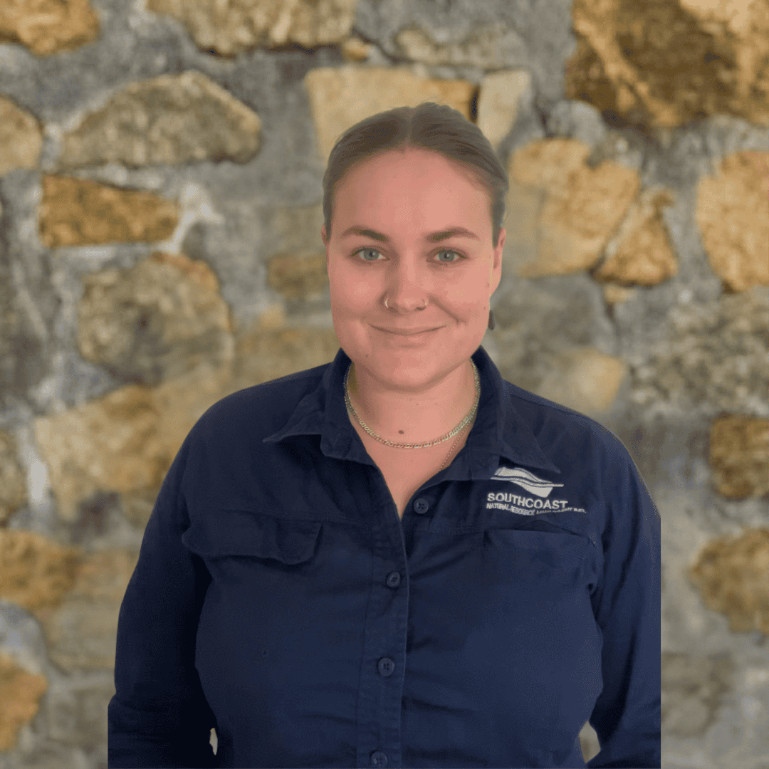 Jayme A person in a navy shirt stands in front of a stone wall, smiling at the camera.