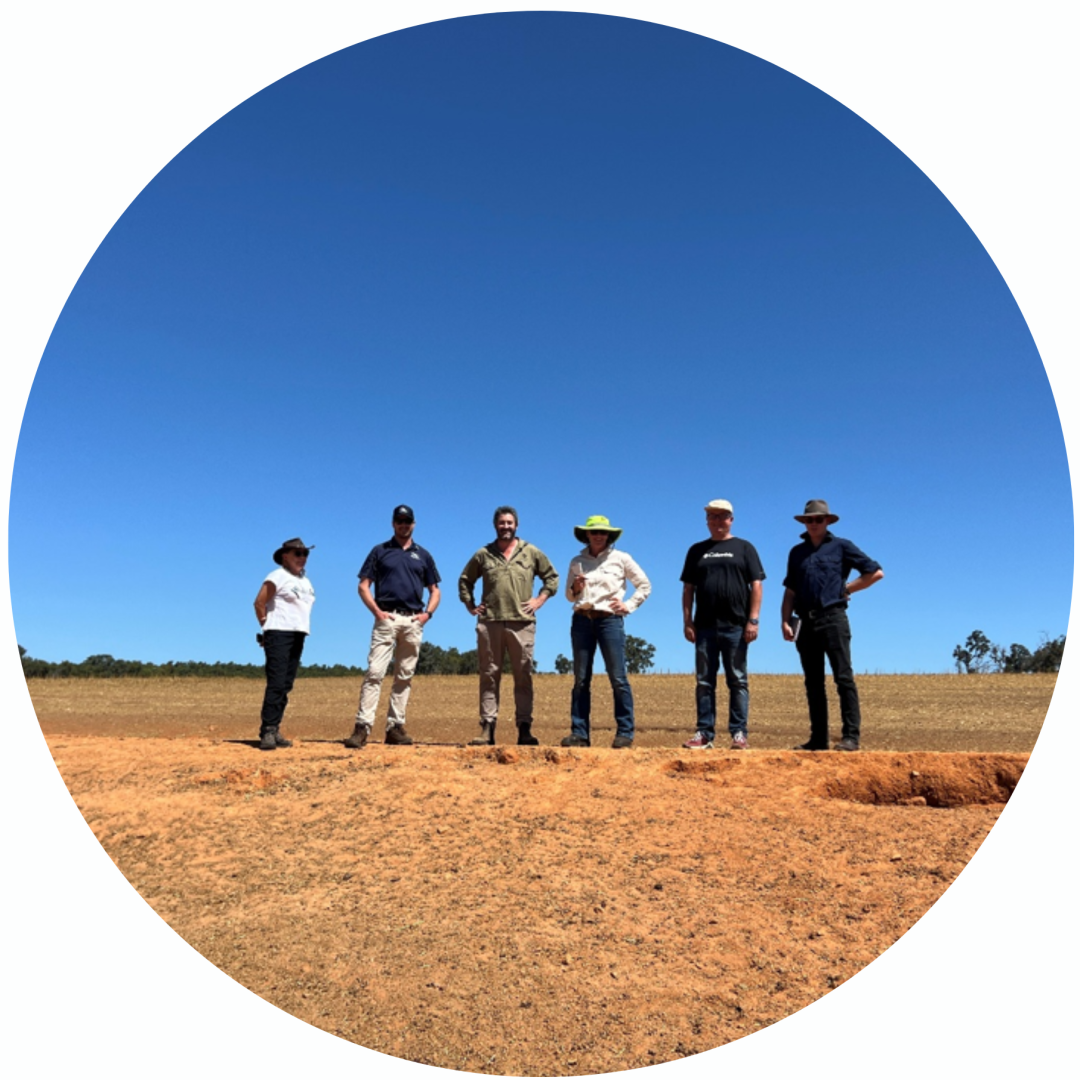 Six people stand on dry, reddish ground under a clear blue sky in an open, rural landscape.