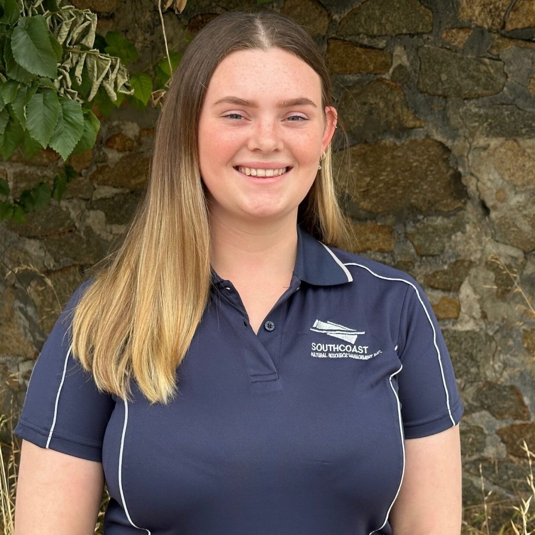 Renae Smiling woman in a navy polo shirt stands outside in front of a stone wall and a tree.
