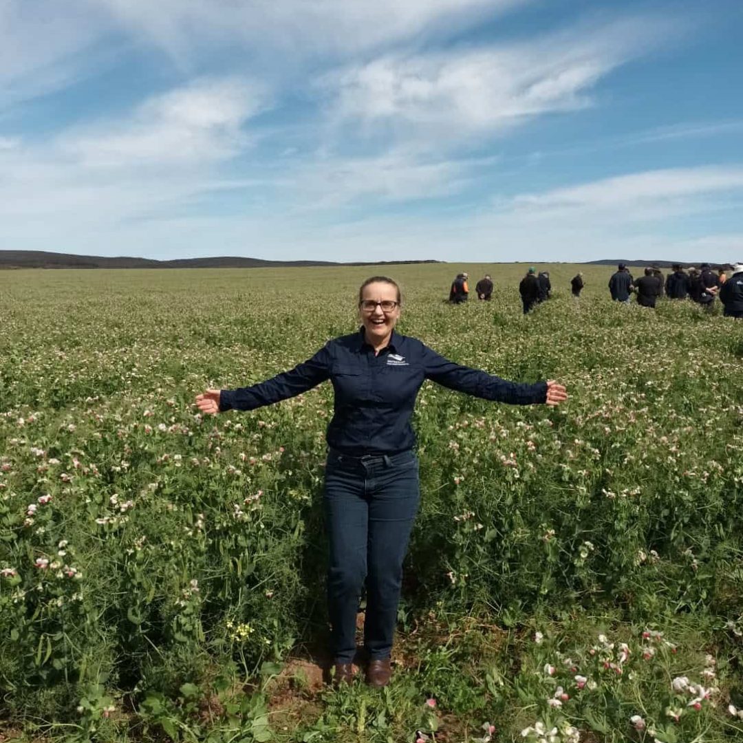 A person stands smiling with arms outstretched in a field of green plants under a blue sky with clouds.
